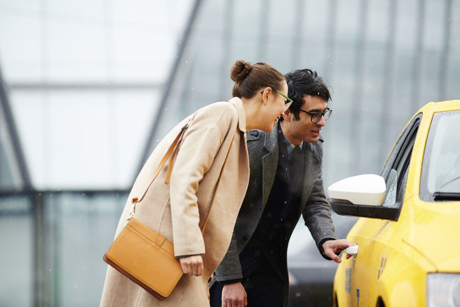 A female passenger in a beige coat and a male taxi driver in glasses both smiling and looking towards the yellow taxi car, representing an airport drop passenger interaction for the Taxi eSIM Affiliate Program.