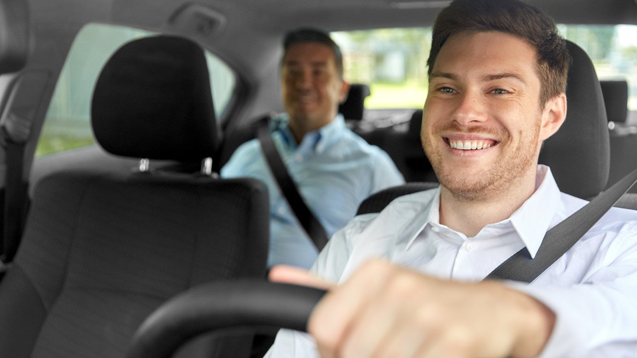 A smiling taxi driver, wearing a white shirt and seatbelt, talks with a blurred passenger in the back seat, illustrating how to talk to airport drop and pickup customers about eSIMs for the Taxi eSIM Affiliate Program.