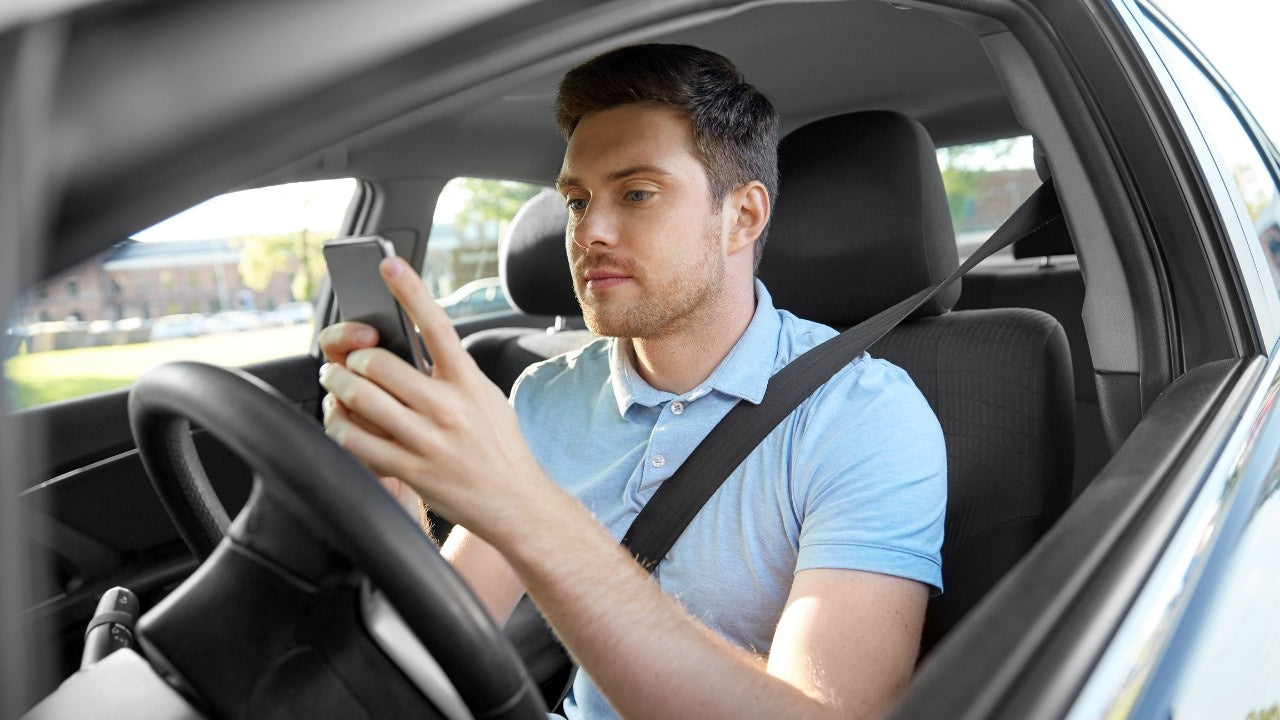 
A taxi driver sitting in his car, wearing a light blue polo shirt and seatbelt, looking attentively at his smartphone, representing checking email for the Taxi eSIM Affiliate Program.