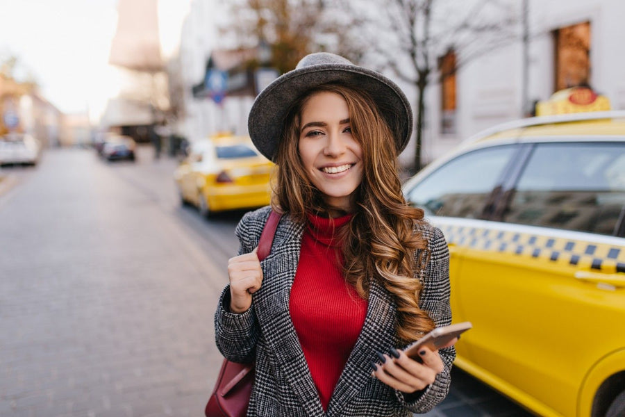 
A smiling female traveler wearing a hat and carrying a bag, holding a smartphone, with yellow taxis visible in the background on a city street, representing an airport pickup scenario for the Taxi eSIM Affiliate Program.